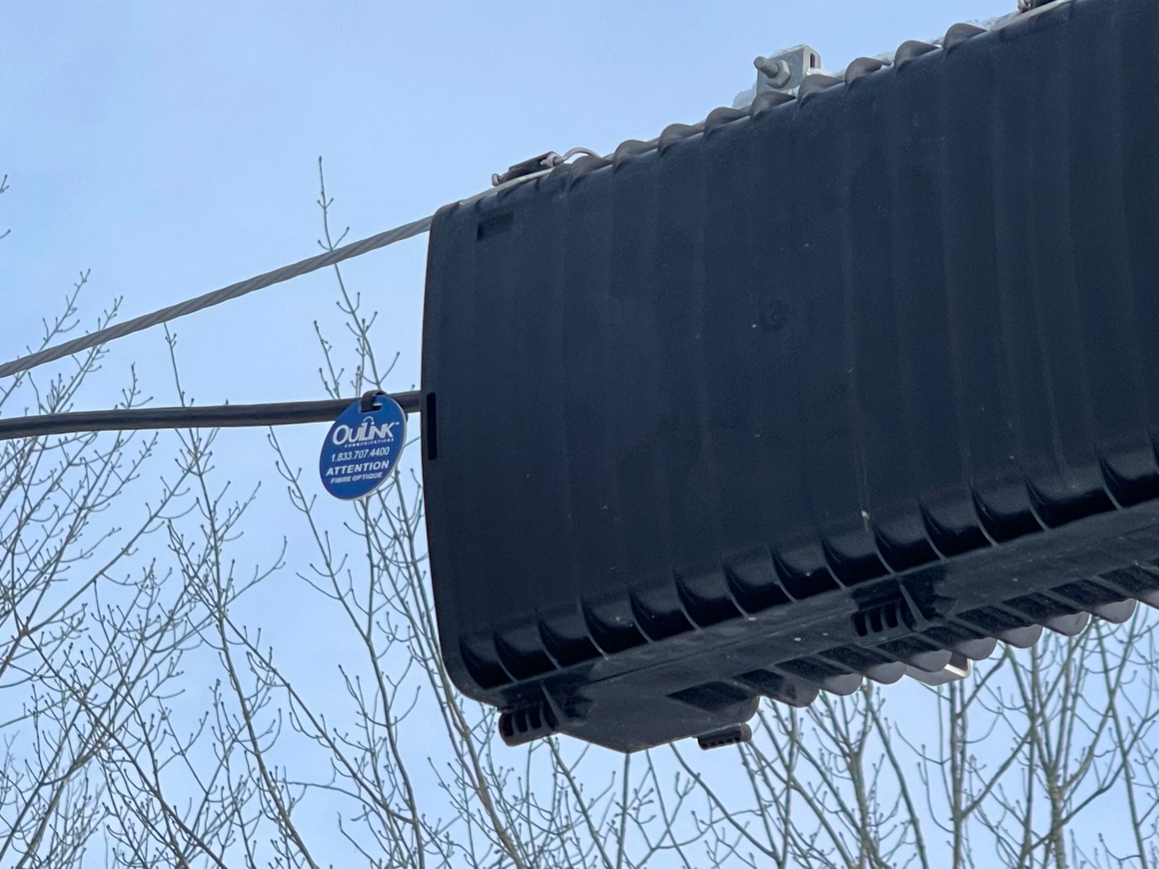 Black ribbed fiber optic splice closure hanging from utility wires with a blue tag.