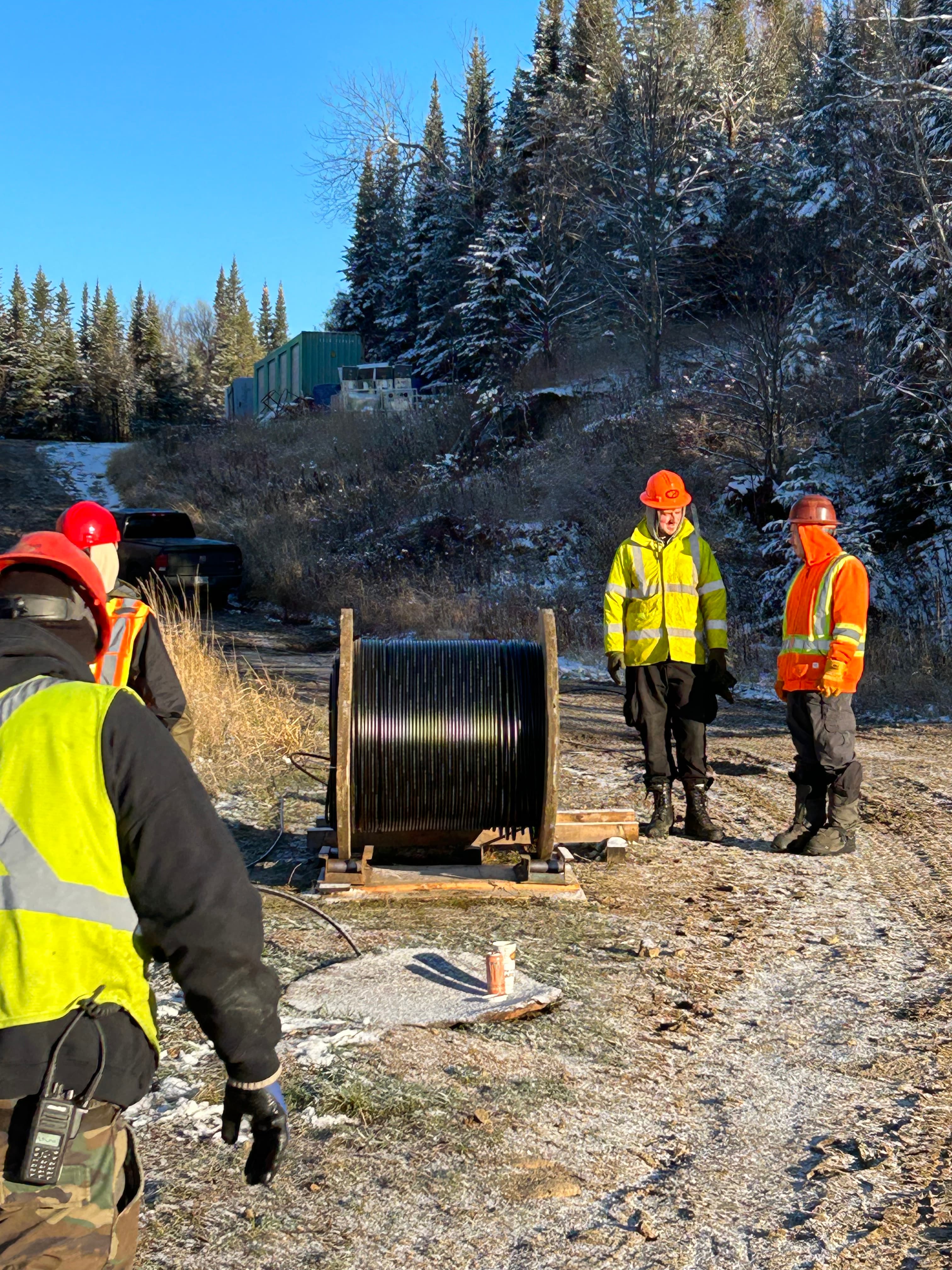 Workers in high-visibility gear stand near a large cable spool in a frosty landscape.