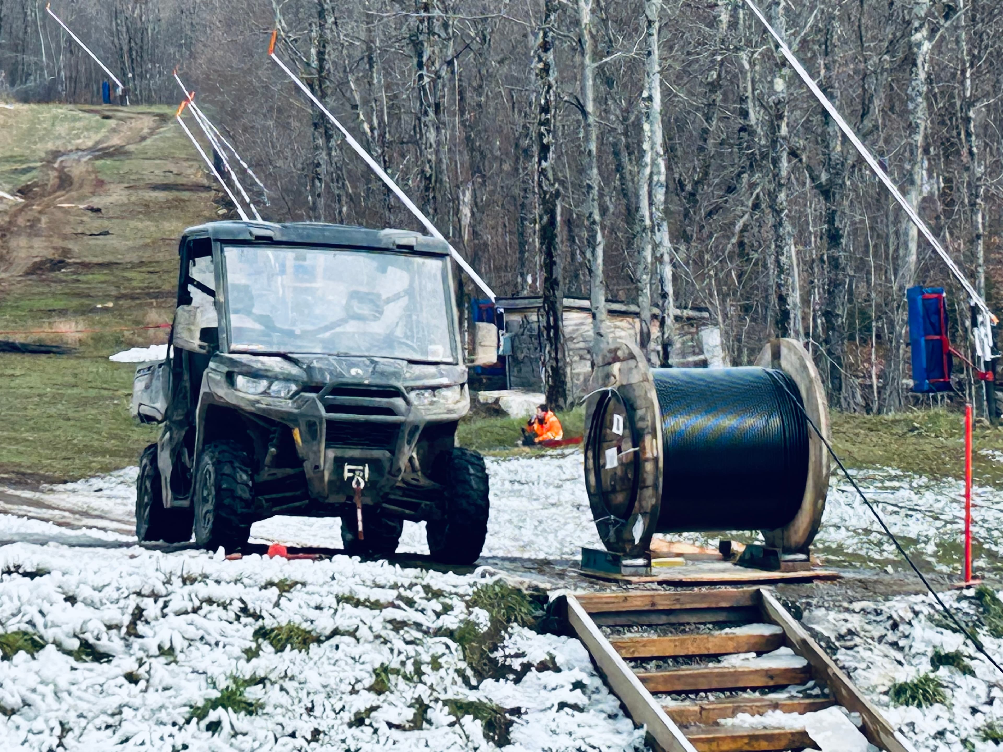 UTV parked beside a large cable spool on a snowy, wooded hillside near power lines.