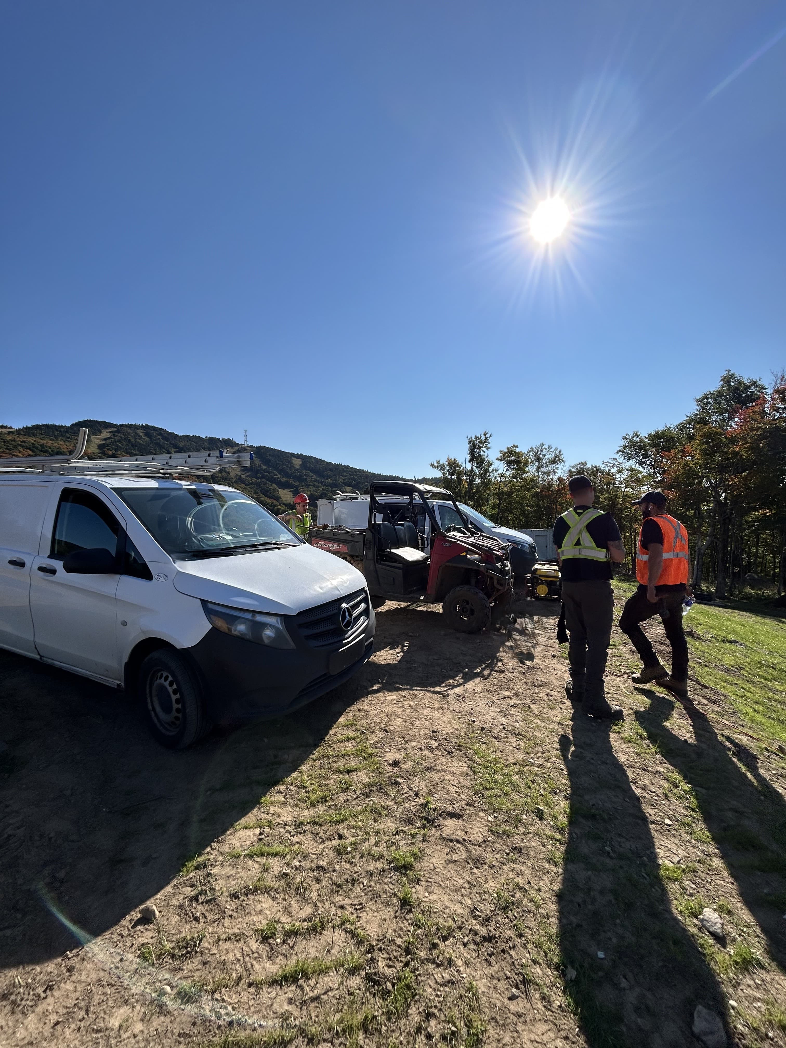 Workers in safety vests stand near a white van and red UTV under bright sunlight.